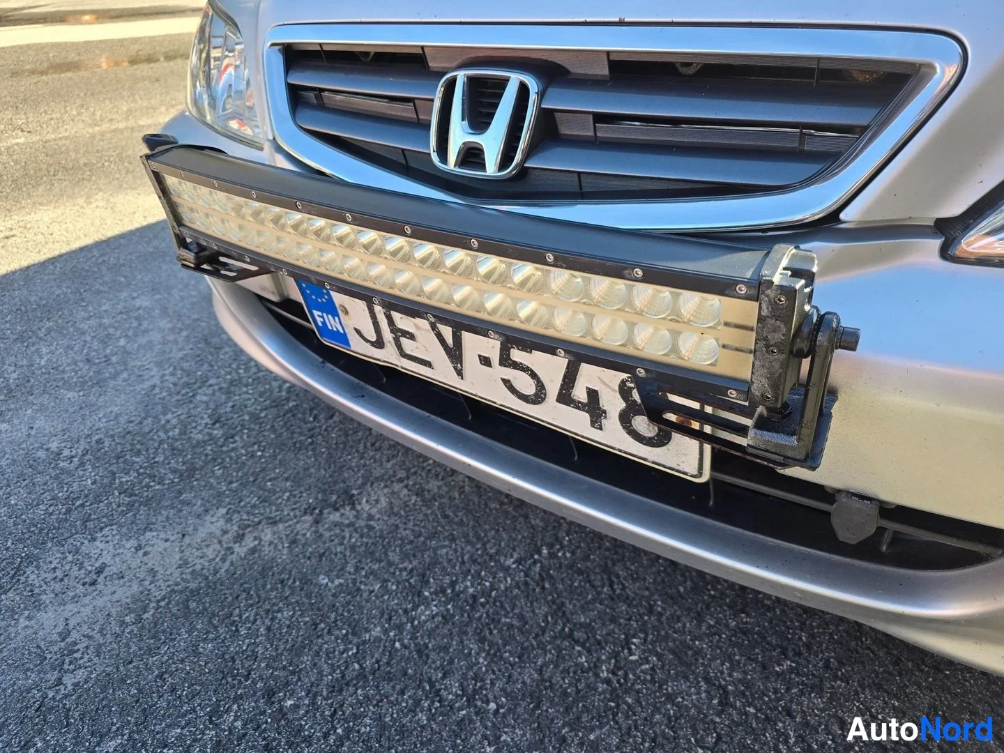 Edestä: Hopeisen Honda-automerkin etunosa lähikuvassa: etusäleikkö ja H-merkki, etupuskurin editse kiinnitetty pitkä LED-työvalo, rekisterikilpi.