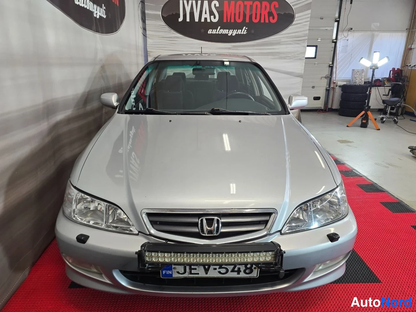 Edestä: Hopeanvärinen Honda Accord -sedan suoraan edestä nähtynä; etusäleikkö Honda-merkillä, ajovalot, puskurissa kiinnitetty LED-lisävalo ja -rekisterikilpi.
