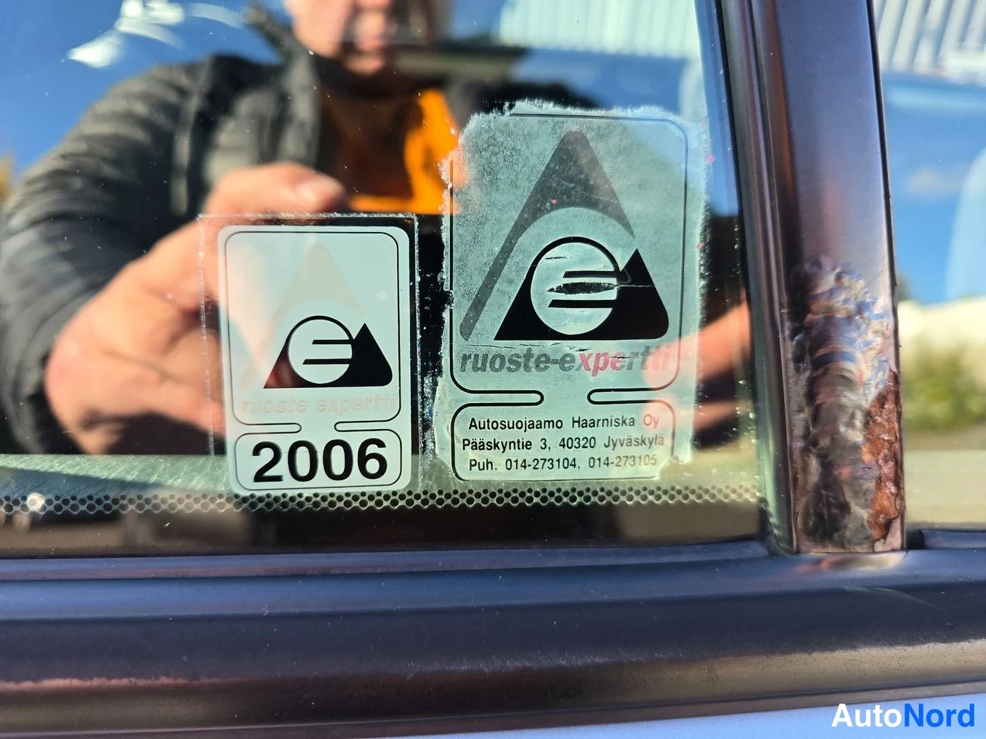 Yksityiskohta: Tuulilasin tarrat: ruoste-expertti-tarra vuosiluvulla 2006 ja isompi ruoste-expertti-tarra, jossa yritystiedot Autosuojaamo Haarniska Oy.