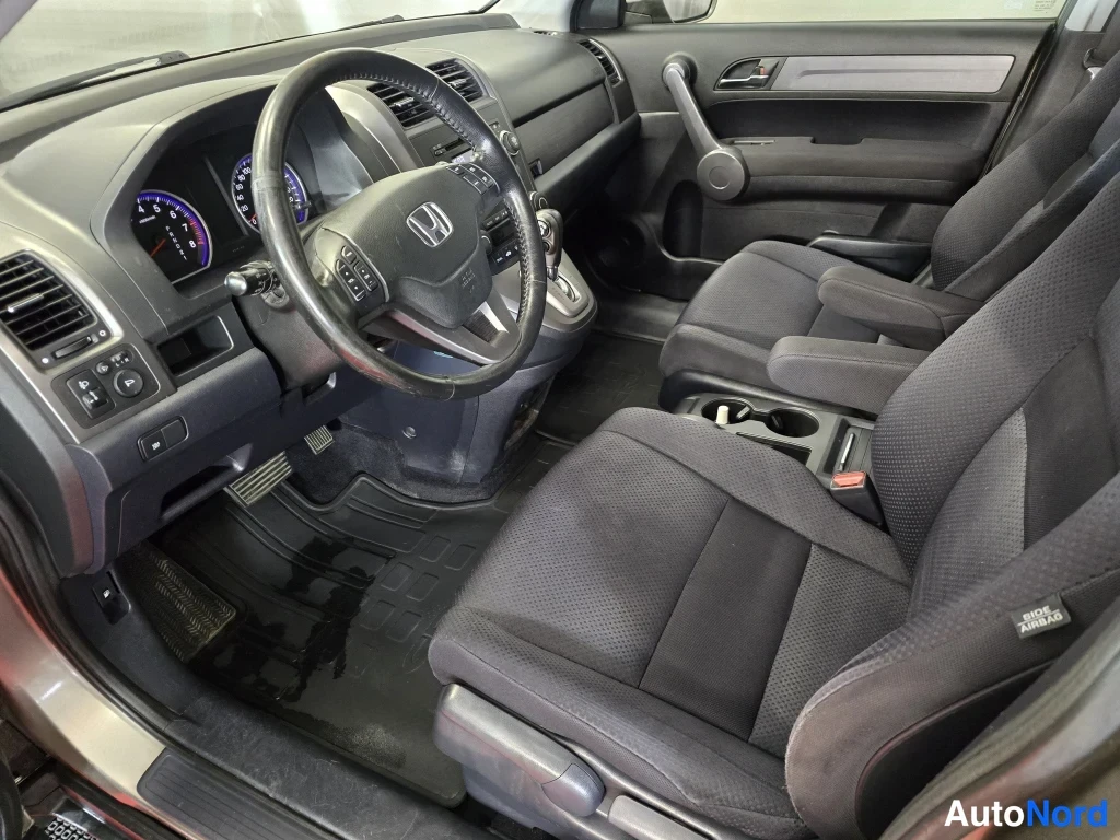 Sivulta: Honda CR-V:n etuistuinverstas tummalla verhoilulla: ohjauspyörä Honda-merkillä, analogiset mittarit, manuaalivaihde seisoo P-asennossa ja painikkeineen, sähköik
