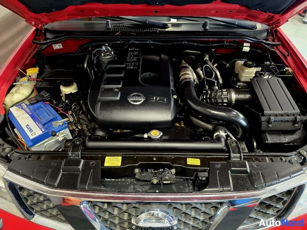 Muu: Punaisen Nissanin avattu moottoritila, Nissan-merkitty moottorin kansi, Varta-akku ja ilmaputket näkyvissä.