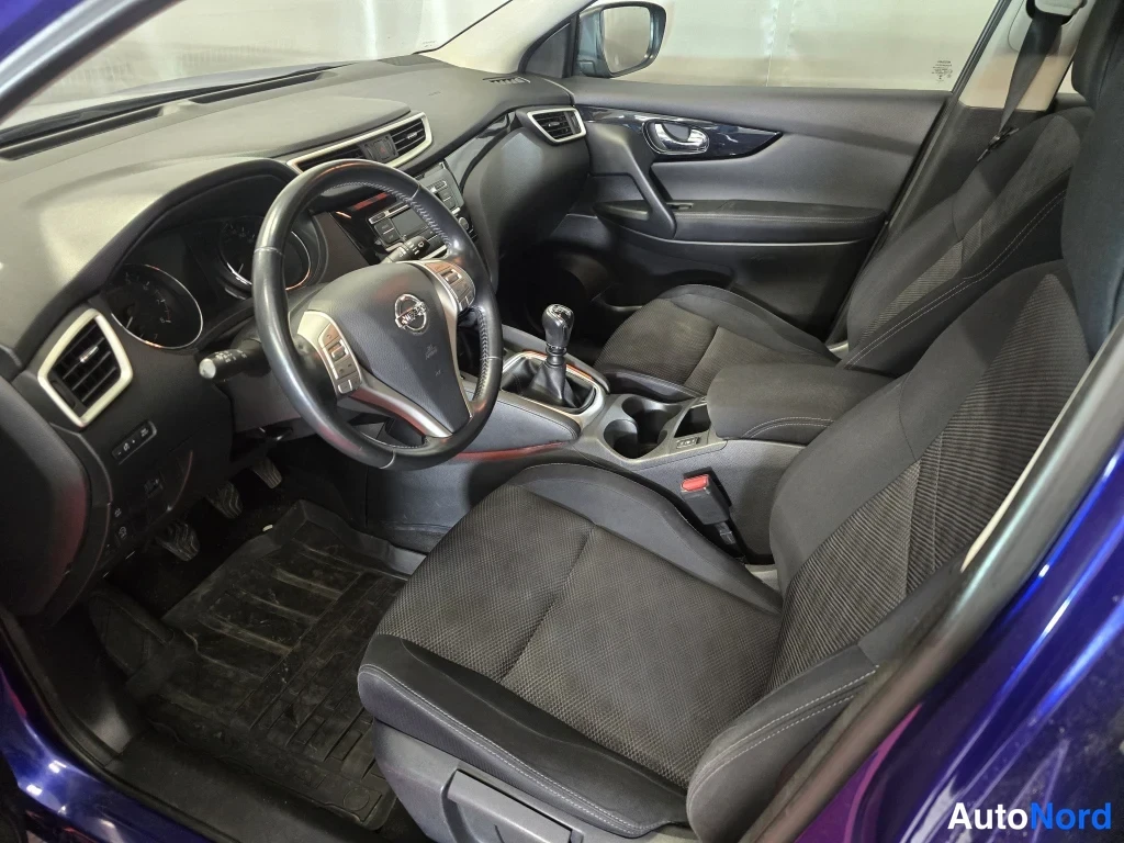 Edestä: Nissan Qashqain matkustamo etumatkustajan puolelta: manuaalivaihteiston vaihdekeppi, kaksipuolinen ohjauspyörä Nissan-logolla ja tummat kangasistuimet.