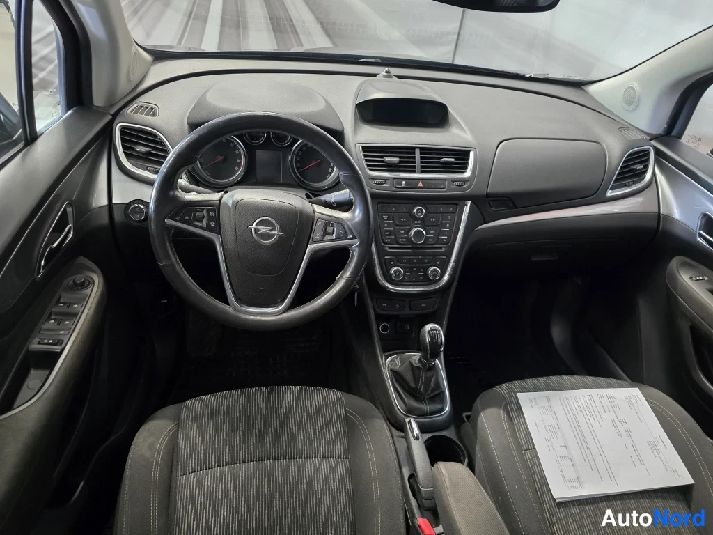 Muu: Opel Mokkan sisätilat etupenkiltä: ohjauspyörä Opel-emblemillä, mittaristo, keskikonsoli ja vaihdevipu, istuimella paperiasiakirja.