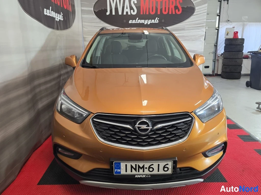 Edestä: Oranssi Opel Mokka X suoraan edestä, etusäleikkö, ajovalot ja rekisterikilpi näkyvät.