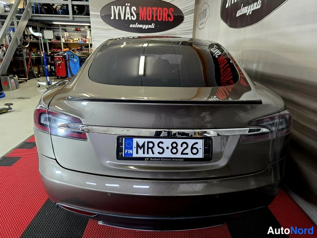Takaa: Tesla Model S -sedan suoraan takaa; rekisterikilpi, vilkkuvalot ja takalasin pinta heijastaa hallin mainostaulua.