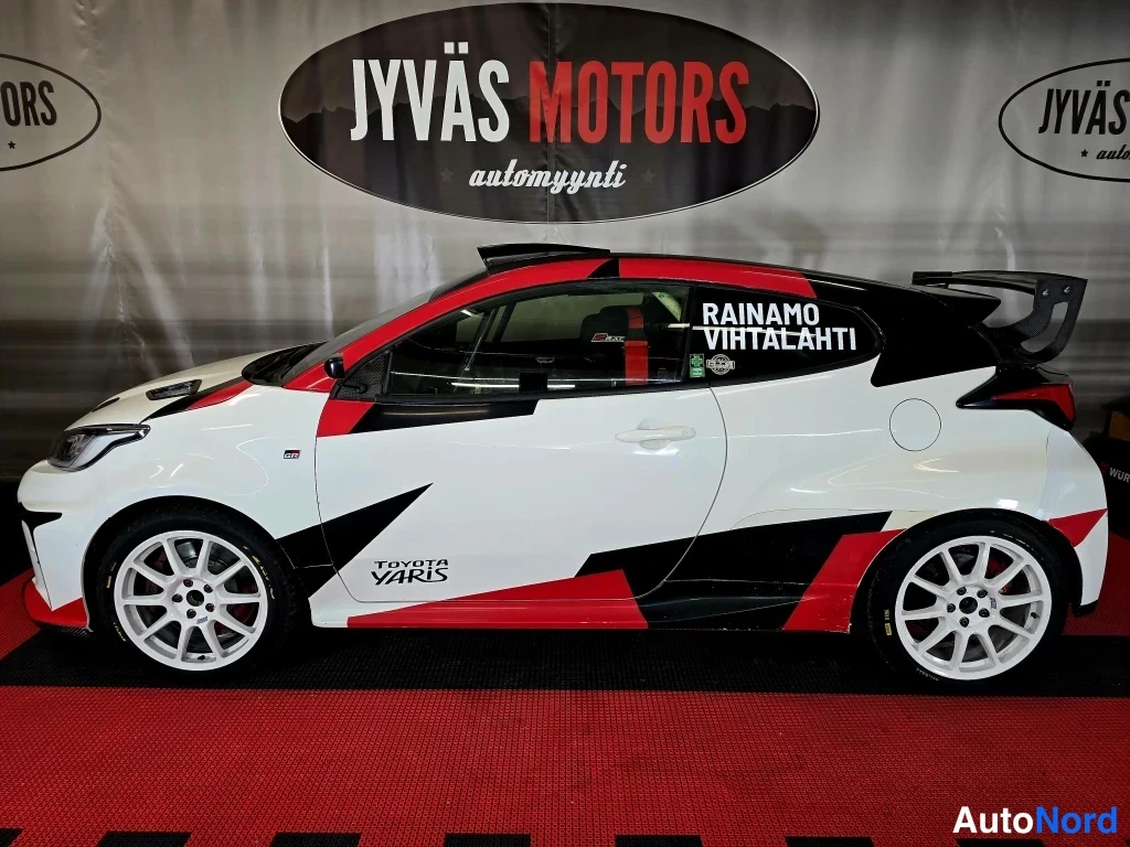 Sivulta: Toyota Yaris -ralliauto sivulta, valkoinen punamustalla grafiikalla, taustalla Jyväs Motors -mainoskangas.
