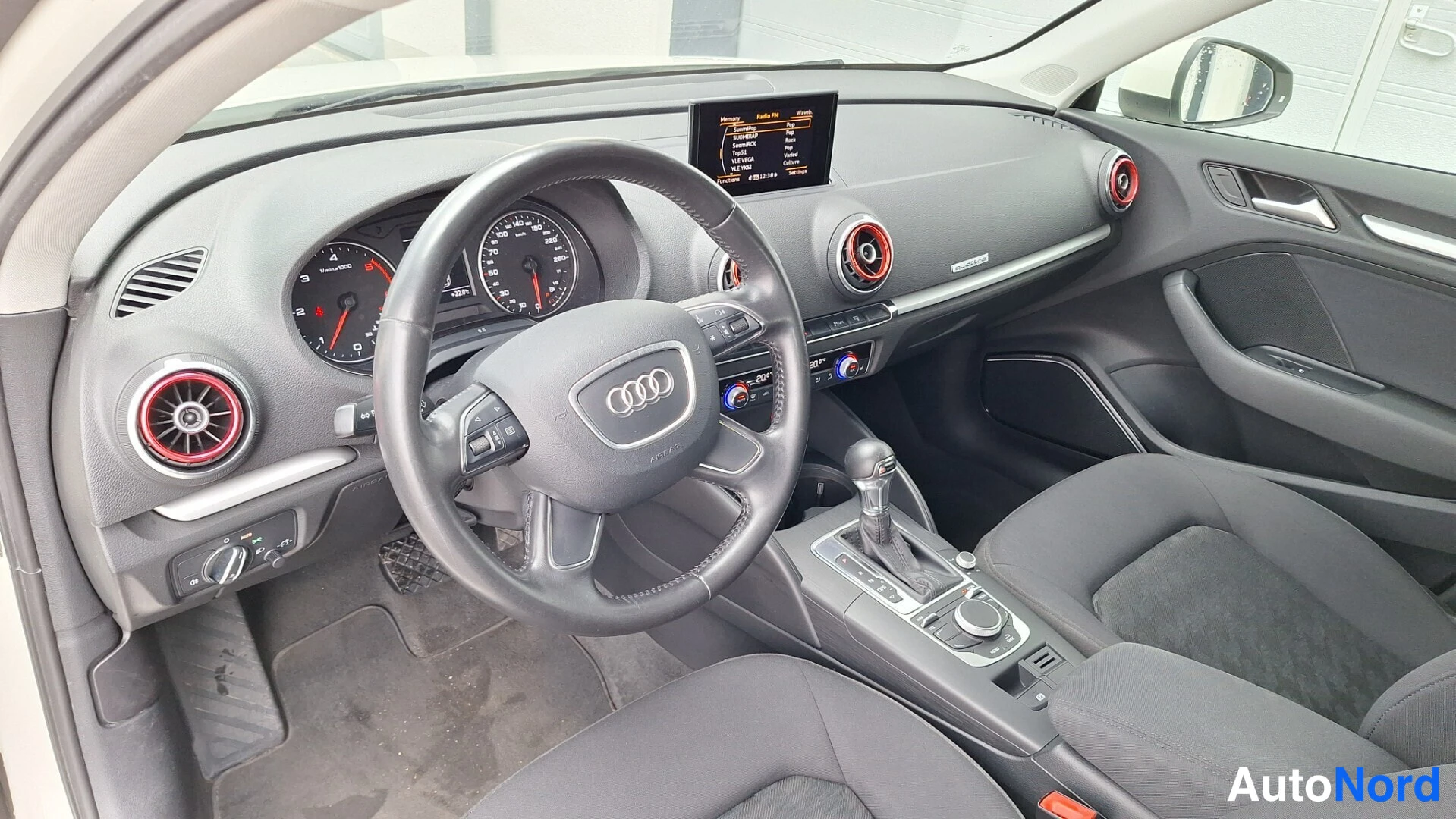 Muu: Kuljettajan puolen sisätila ylhäältä: Audi-ohjauspyörä, mittaristo, ylös kääntynyt keskinäyttö, punareunuksiset ilmastointisuuttimet ja automaattivaihteiston va
