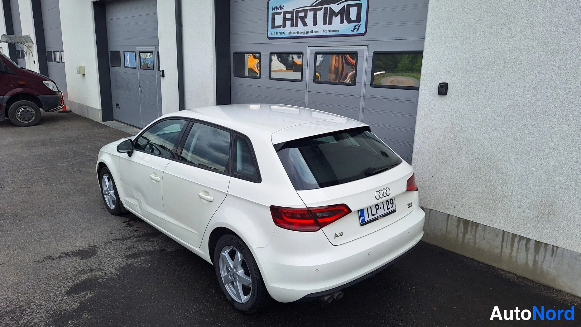 Takaviisto: Valkoinen Audi A3 -viistoperä takaviistosta kuvattuna hallin ovia edessä.
