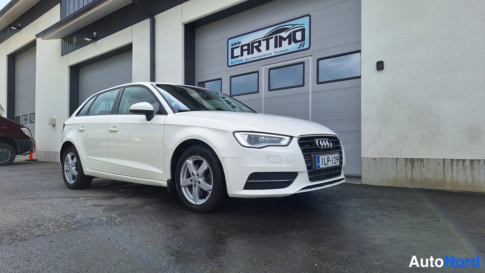 Etuviisto, nokka oikealle: Valkoinen Audi A3 Sportback kolmesta neljäsosasta etuosasta oikealle kuvattuna huoltamorakennuksen edessä.
