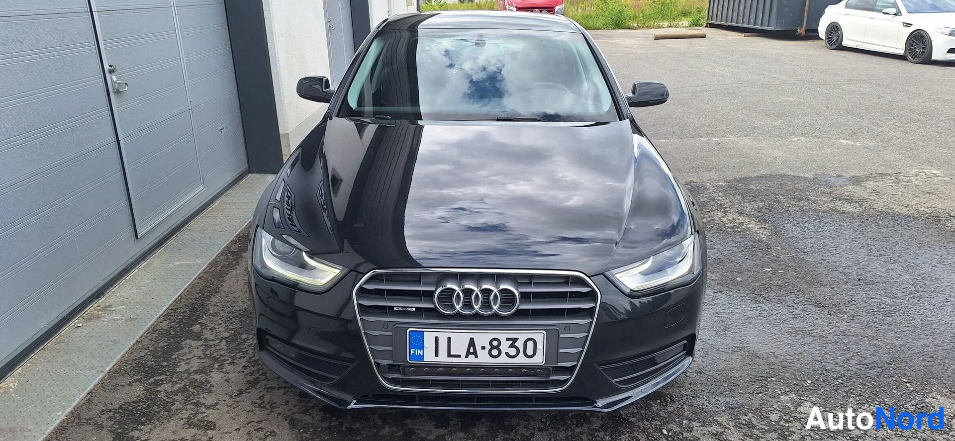 Edestä: Audi A4 -sedanin suora etukuva: musta eturunko, Audi-merkki grillissä ja rekisterikilpi näkyvät. Muut kuvan autot ovat taustalla, eivät arvioitava kohde.