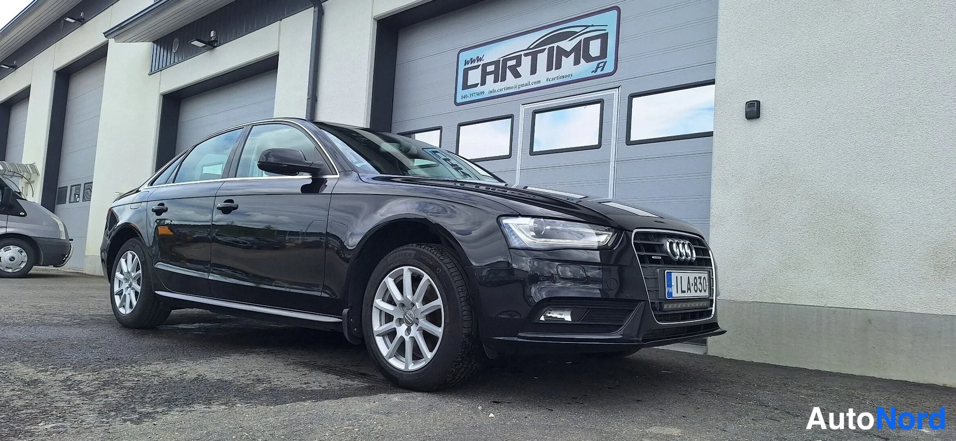 Etuviisto, nokka oikealle: Mustan Audi A4 -sedanin etukulma oikealta, rekisteritunnus, huoltamon ovella.