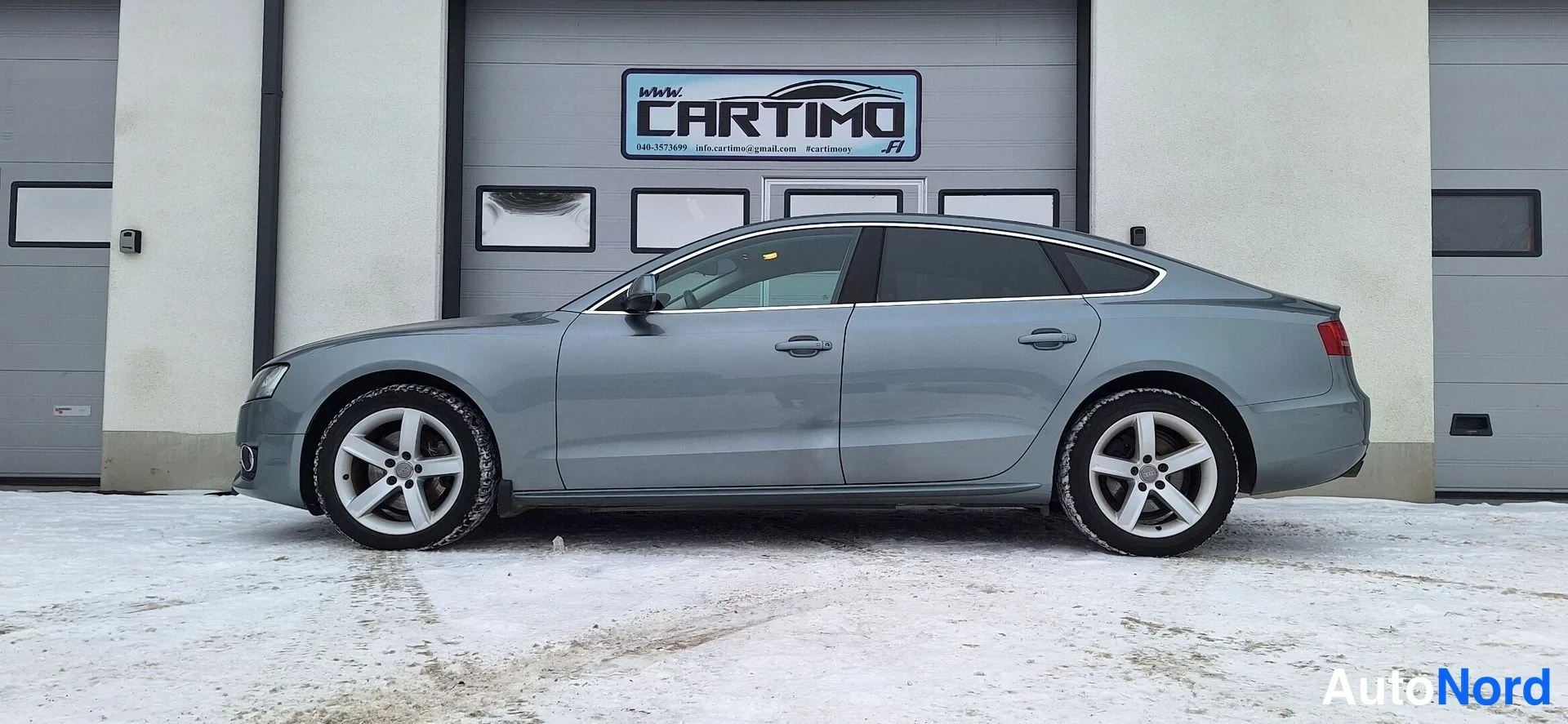 Etuviisto, nokka vasemmalle: Audi A5 Sportback -henkilöauto sivulta, harmaa kori, viisiovinen coupémaiset linjat, talvimaisema lumisella pihalla.