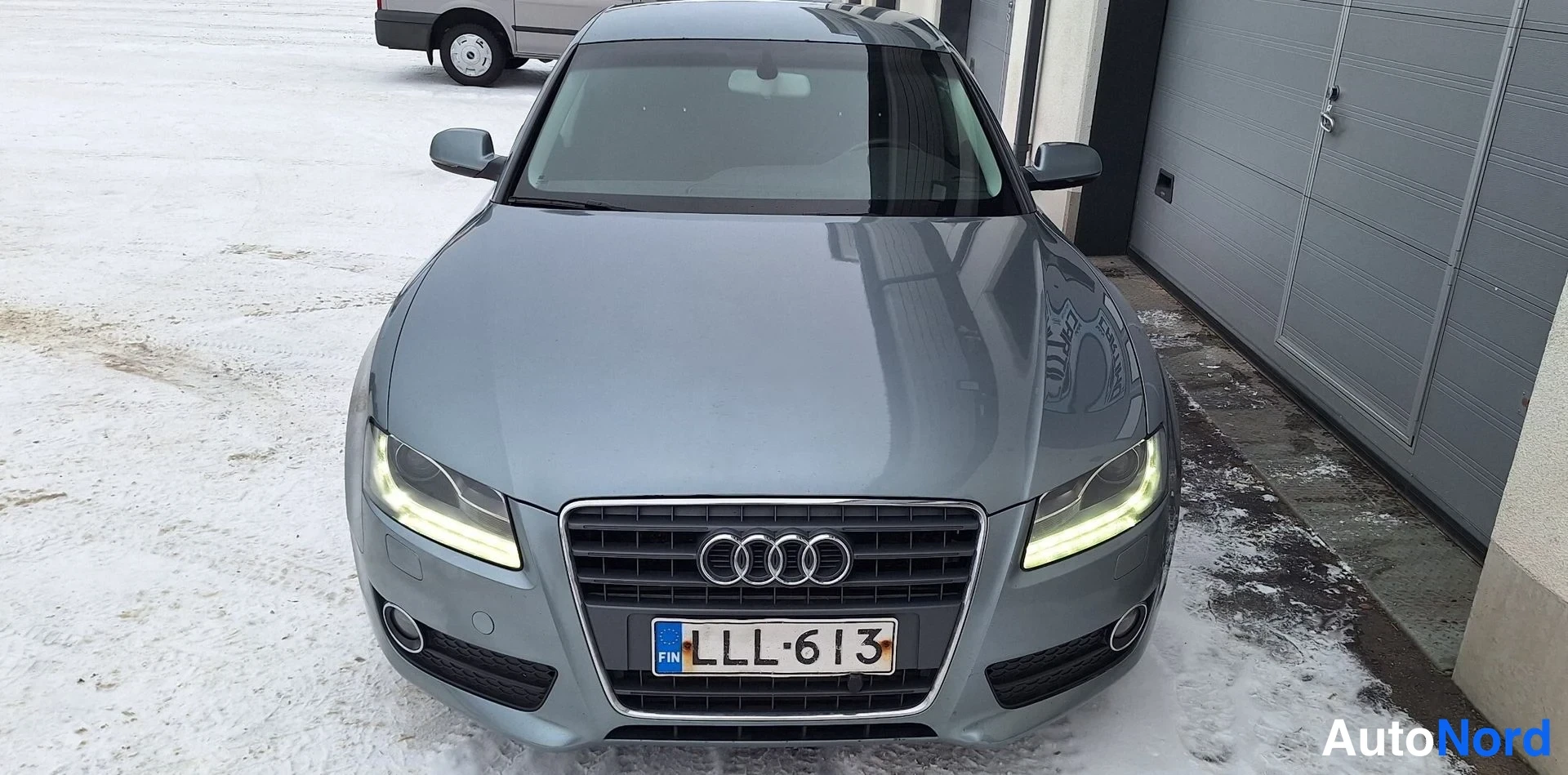 Edestä: Edestä suoraan kuvattu harmaahenkinen hopeanharmaa Audi, jonka etusäleikössä on neljän renkaan merkki ja punakellotauluinen rekisterikilpi.