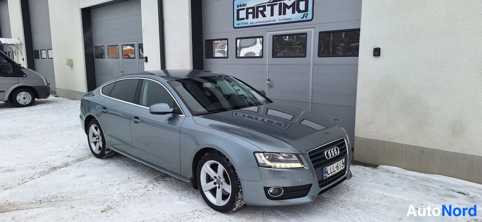 Etuviisto, nokka oikealle: Audi A5 Sportback etukulmasta oikealta, keulakorsi ja kylki näkyvät, harmaa kori, rekisteri LL-613, lunta maassa.