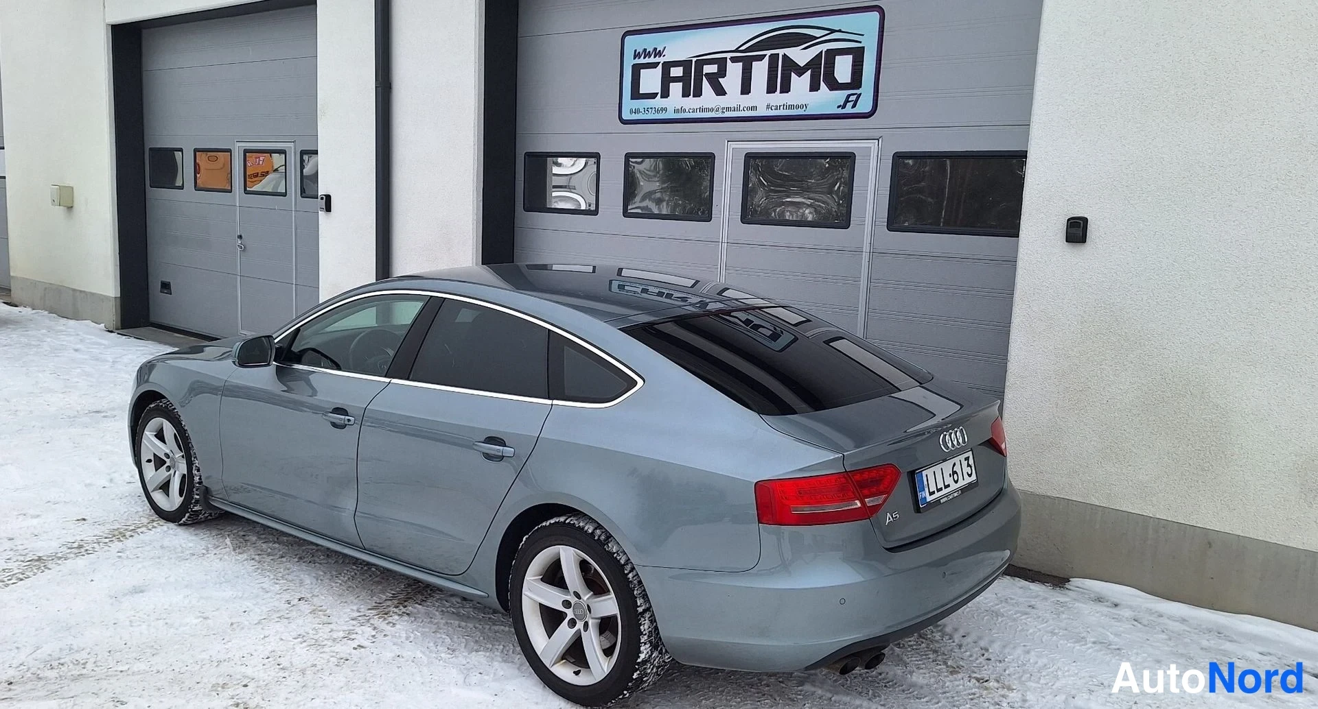 Takaviisto: Audi A5 Sportback takakulmasta, perä ja kylki näkyvät, harmaa kori, punaiset takavalot, rekisteri LL-613, lunta maassa.