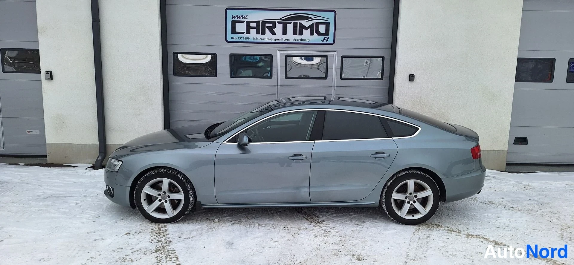 Etuviisto, nokka vasemmalle: Audi A5 Sportbackin kylki ja etuosa, keula osoittaa vasemmalle, lumisella pihalla.