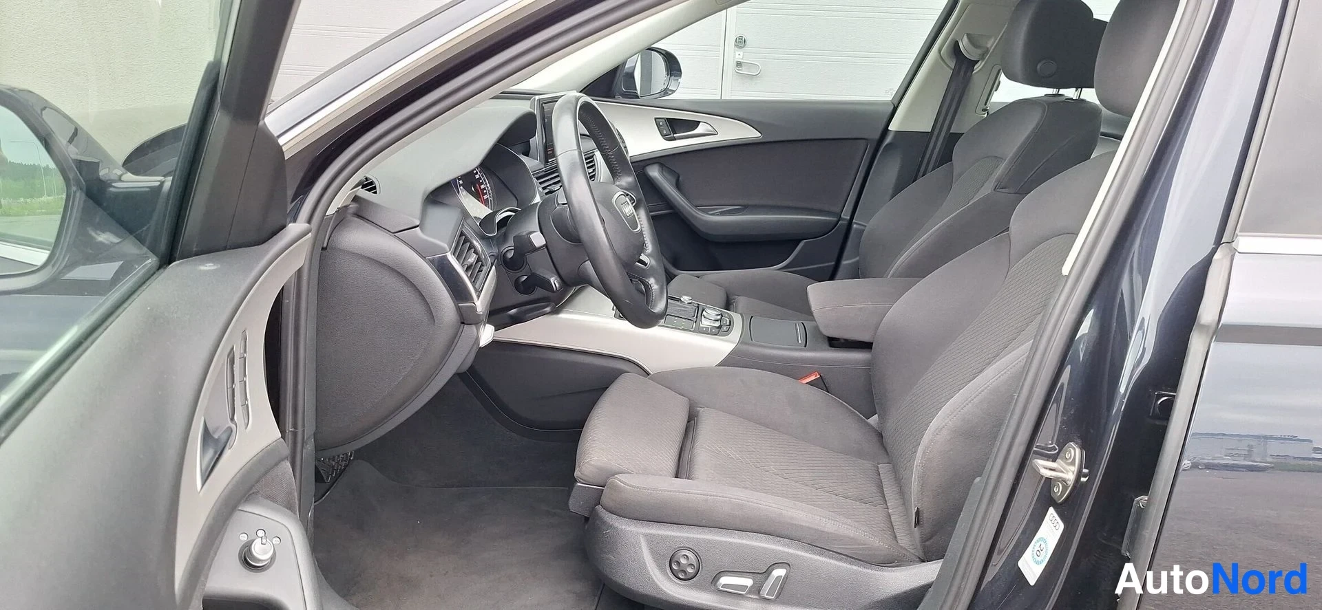 Muu: Kuljettajan ovesta kuvattu ohjaamo: Audi-ohjauspyörä, etuistuimet ja keskikonsoli näkyvät, oven sisäpaneeli vasemmalla.