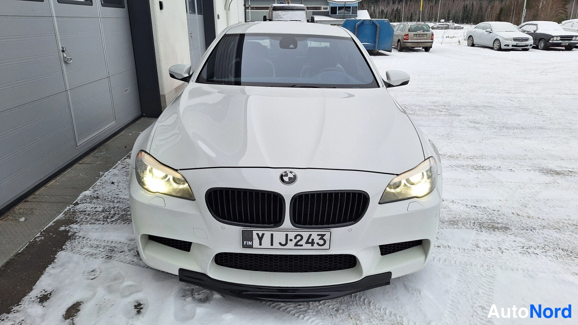 Edestä: Valkoisen BMW-sedanin etukuva lumisessa pihalla, etuvalot päällä, rekisterikilpi.