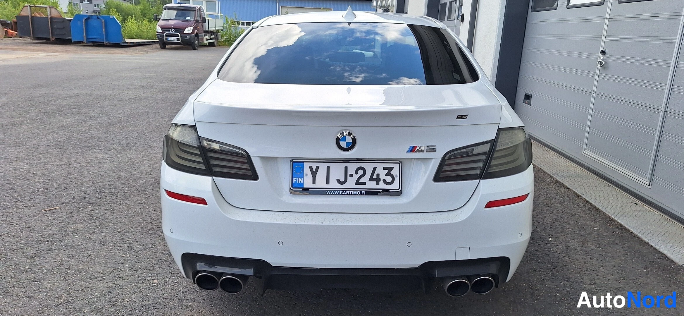 Takaa: Valkoisen BMW-sedan takaosa suoraan takaa, M5-merkki, tummat takavalot, neljä pakoputkea, rekisteri.