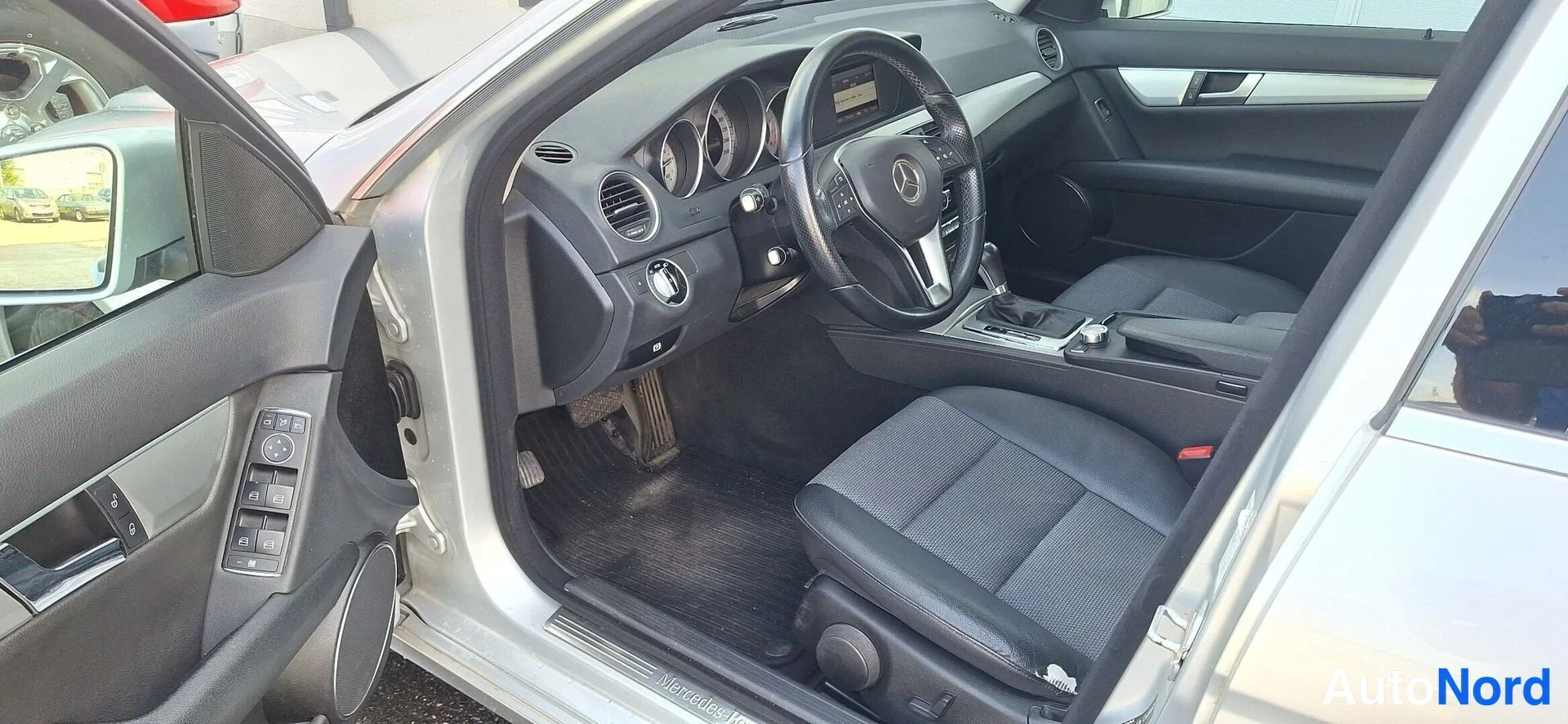 Muu: Sisätilakuva Mercedes-Benz C-sarjan etuosasta: kuljettajan ovi auki, tumma mittaristo, Mercedes-logollinen ohjauspyörä, kangas-penkit ja keskikonsoli näkyvät.