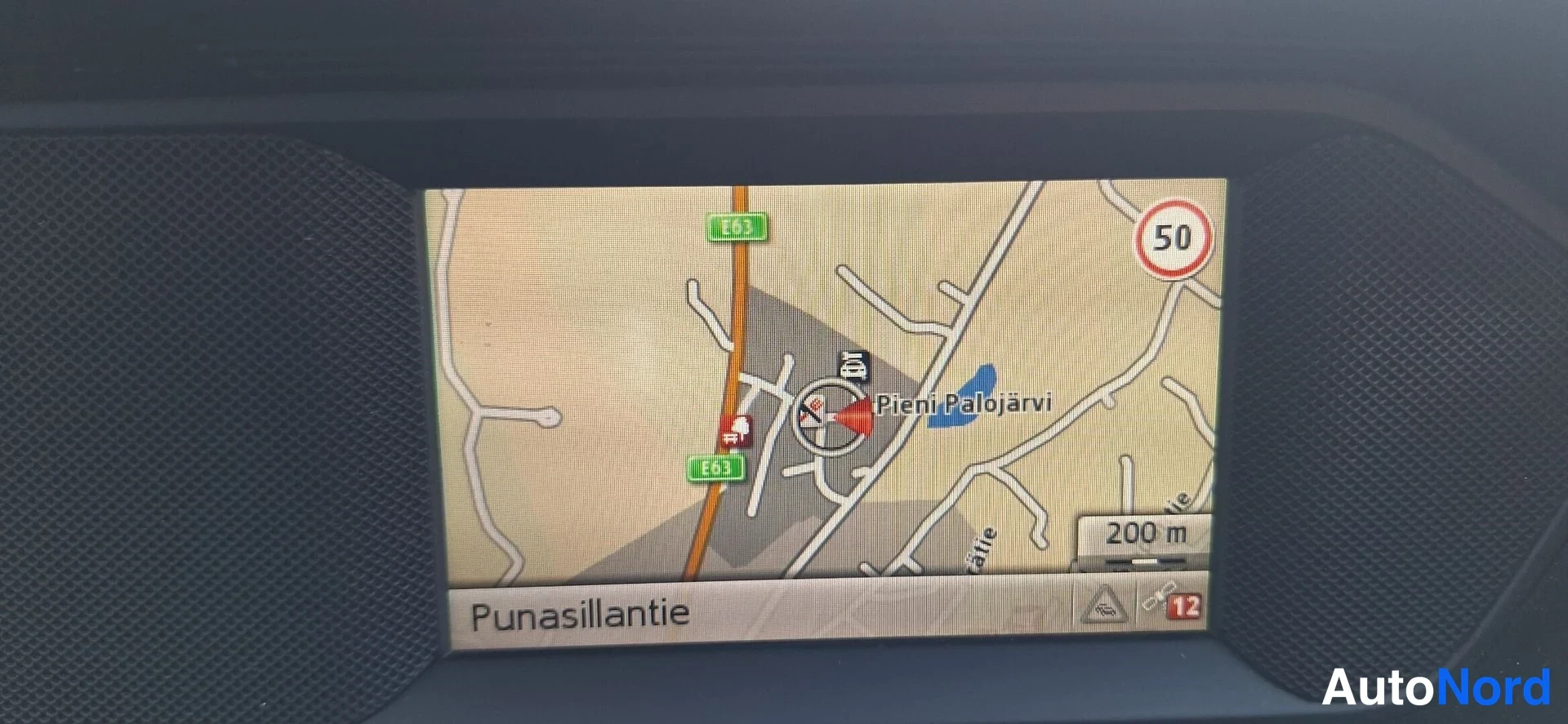 Muu: Kojelaudan näytöltä näkyy navigaattorin kartta, jolla lukee Pieni-Palojärvi, Punasillantie, E63, 200 m ja nopeusrajoitus 50.