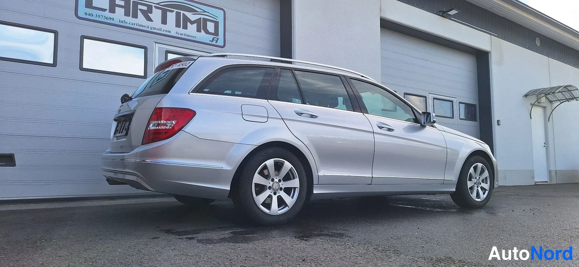 Takaviisto: Hopeinen farmariauto takaa vasemmalta viistosti nähtynä hallin edessä.