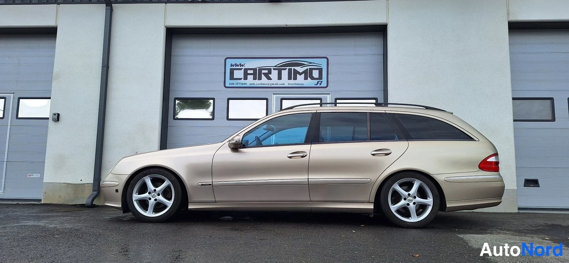 Sivulta: Beige Mercedes-Benz farmari sivulta katsottuna talon seinän edessä.