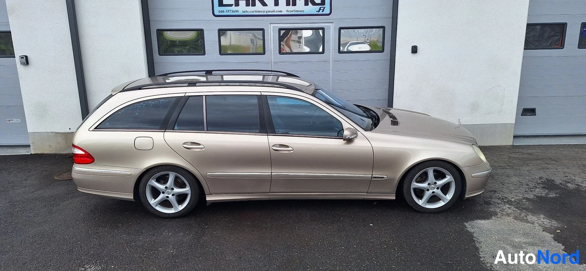 Etuviisto, nokka oikealle: Beige Mercedes-Benz farmari koko sivulta ja etunurkalta kuvattuna; näkyvissä kylki, etuvalo, kattokaiteet ja hopeiset kevytmetallivanteet.
