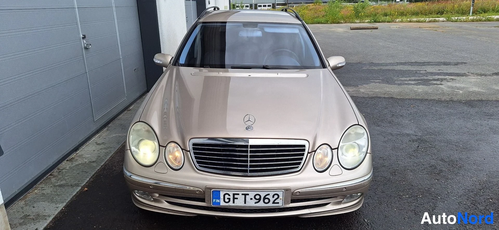 Edestä: Beige Mercedes-Benz farmari suoraan edestä kuvattuna; näkyvissä Mercedes-tähti, jäähdyttjärityyppinen etusäleikkö, pyöreät etuvalot ja rekisteritunnus.
