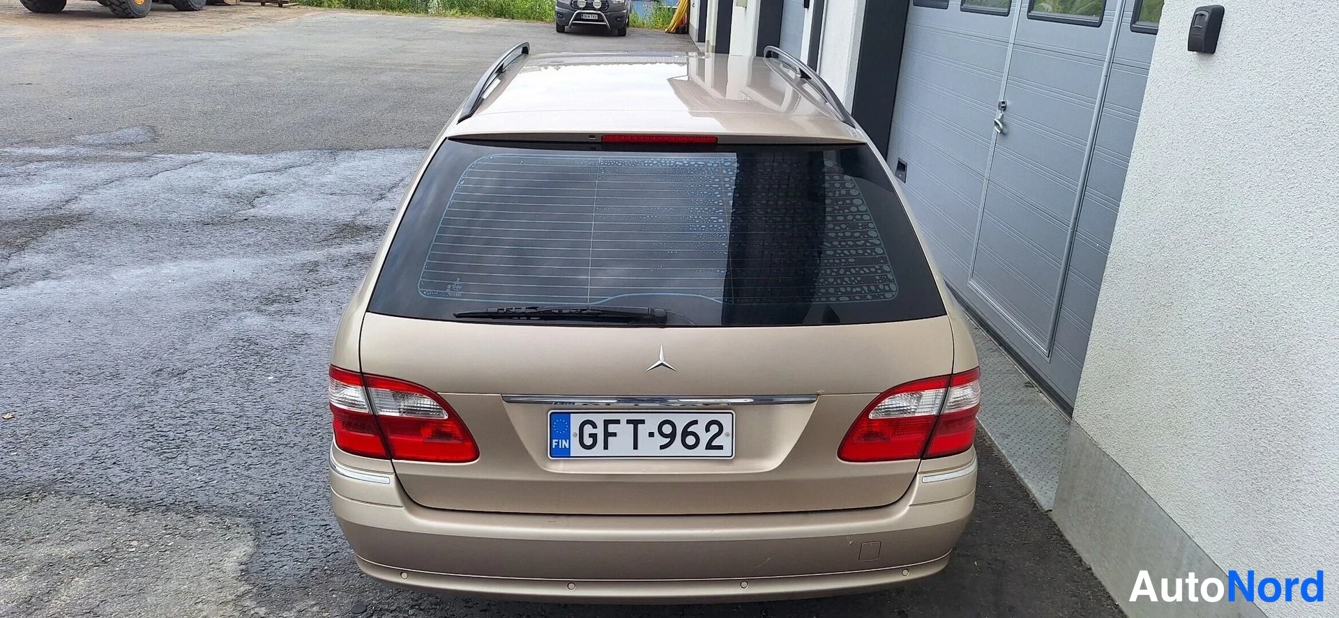 Takaa: Beige Mercedes-Benz -farmariauto takaa päin kuvattuna; takalasin lämmityslinjat, kattotelineet, punaiset takavalot ja rekisterikilpi näkyvissä.