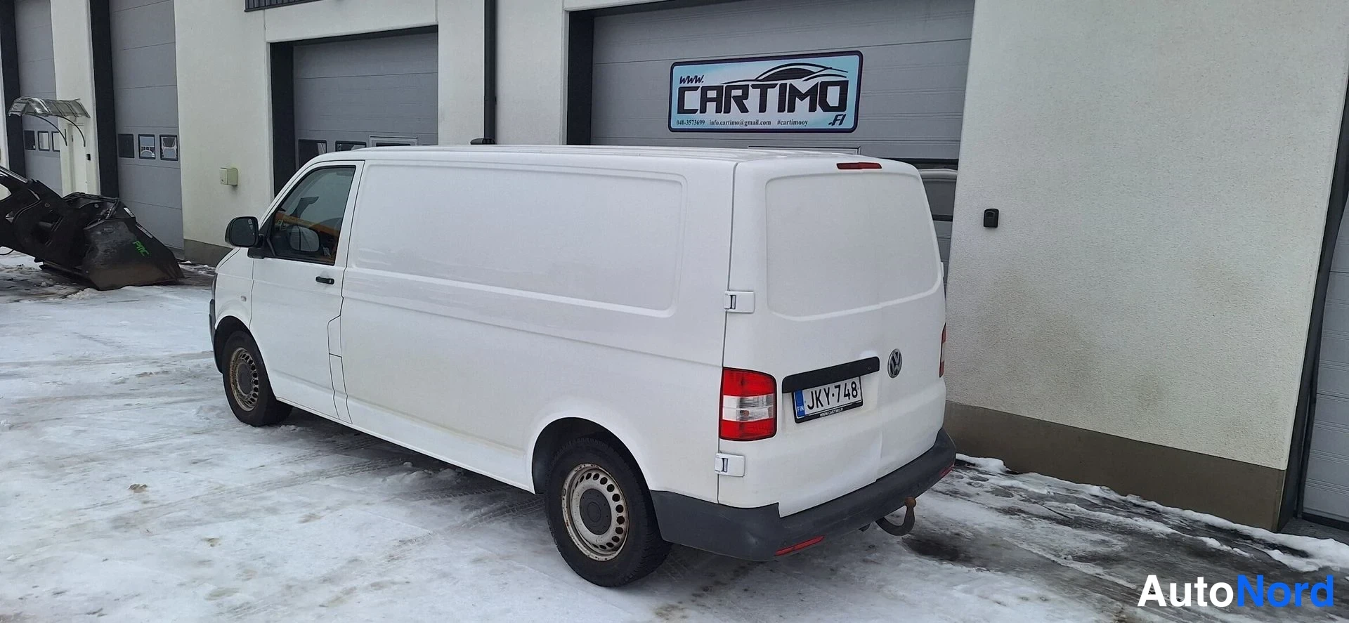Takaviisto: Valkoinen pakettiauto takaa kulmasta talvisella pihalla huoltamorakennuksen edessä.