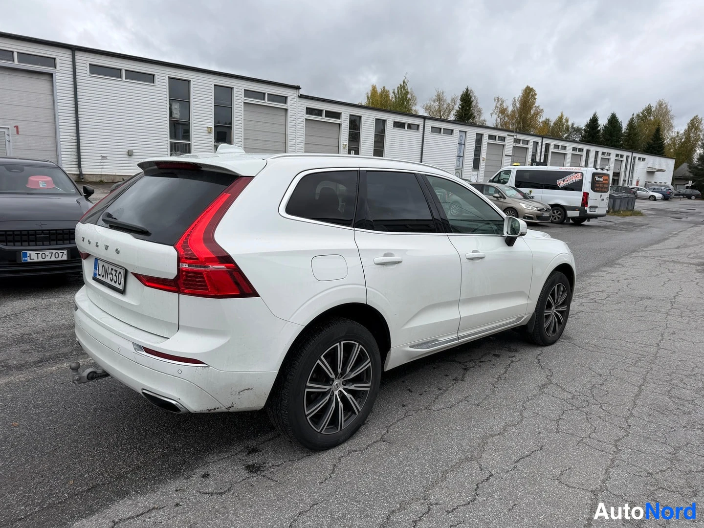 Takaviisto: Valkoisen tila-auton takasivukulma: takapää ja kylki näkyvissä muissa autoissa pysäköidulla pihalla.