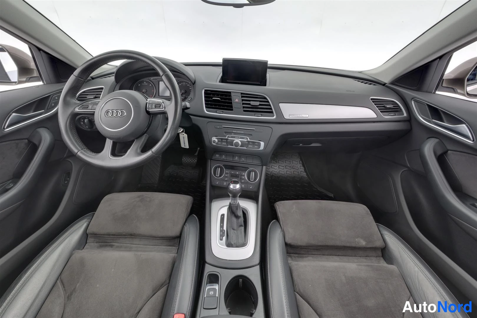 Muu: Audi Q3:n sisätilat: tumma mittaristo ja ohjauspyörä Audi-merkillä, keskikonsolissa vaihtajanappi, nappiryhmät ja kaksi tummaa etuistuinta.