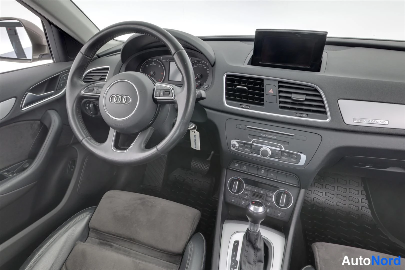 Muu: Audi Q3:n etuistuin ja kojelauta lähikuvassa: tumma nahkainen ohjauspyörä Audi-logolla, keskellä näyttö, Audi median nappirivi ja tummat matot.
