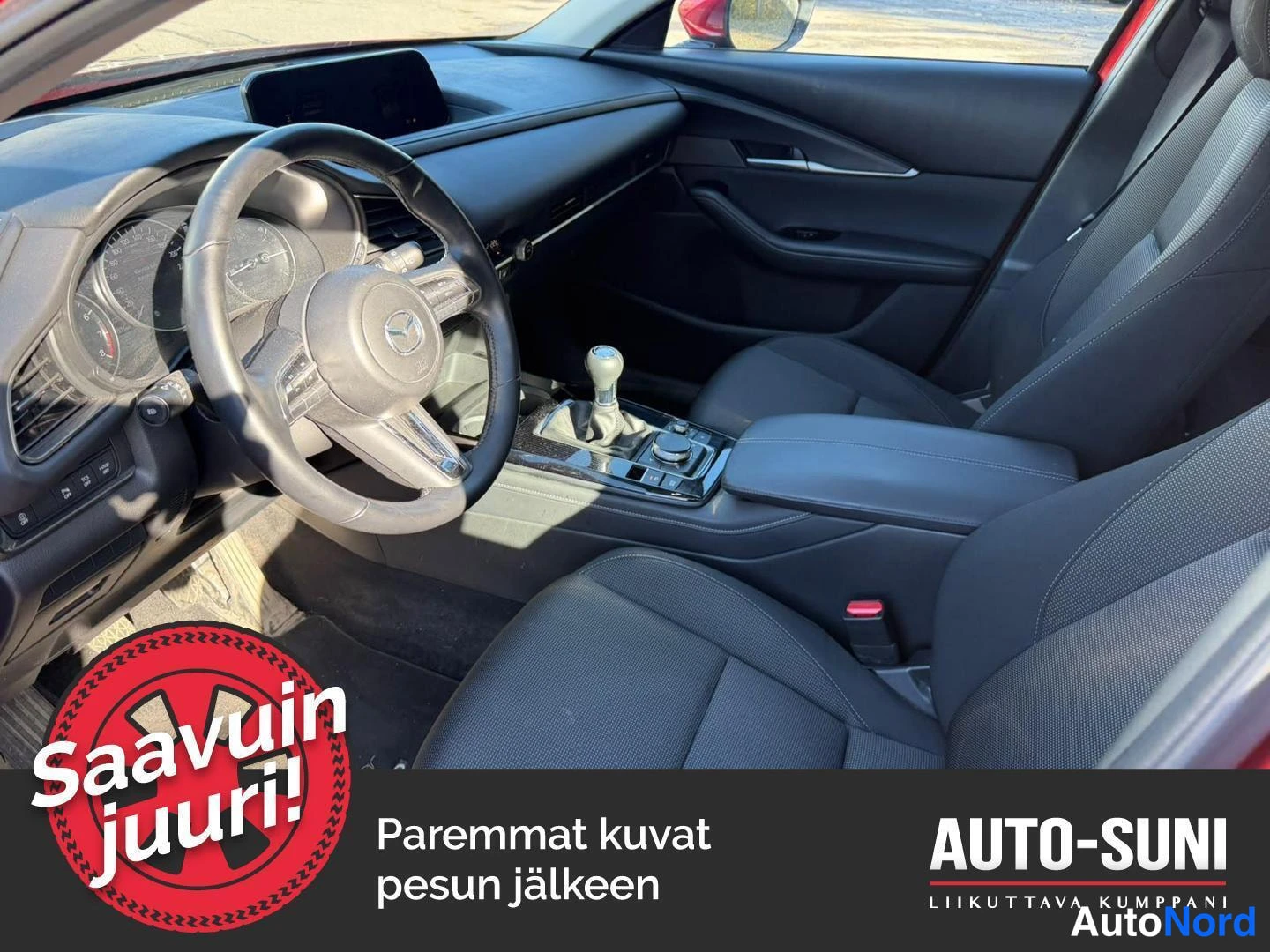 Sivulta: Henkilöauton tumma ohjaamo: Mazda-merkkinen ohjauspyörä, kojelaudan näyttö, keskikonsolin vaihteenvalitsin ja kierto-ohjain sekä kaksi tummaa etuistuinta. Kuvan
