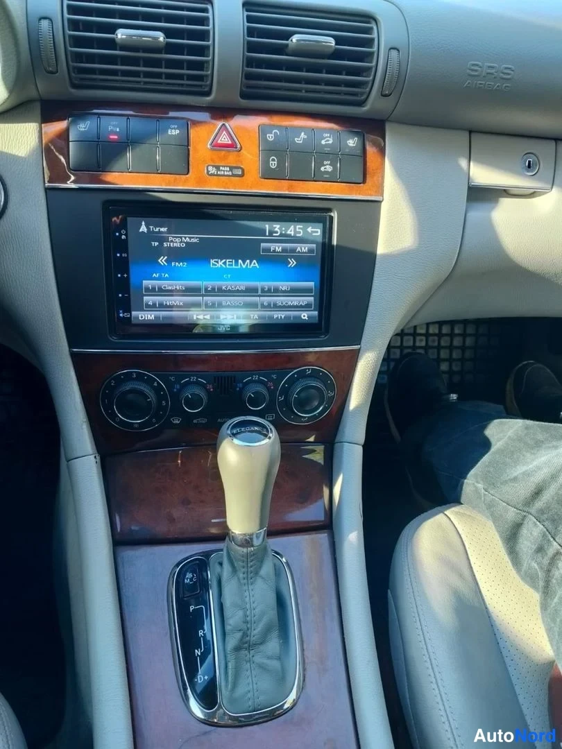 Yksityiskohta: Keskikonsolin ylä- ja välialue automaattivaihteen valitsimella, radio näytöllä teksti SKELMA ja kello 13:45, pukuoristeinen paneli ja ilmastoinnin säätörivit.