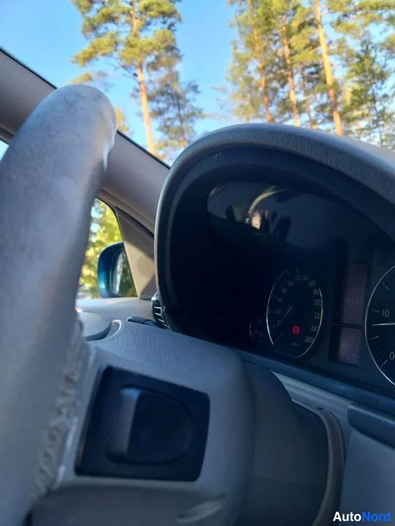Yksityiskohta: Kuljettajan näkyvä alue ohjauspyörä ja osa mittaristosta, ajokortti istuu vasemmalla, taustalla näkyy puistoa ikkunan kautta.