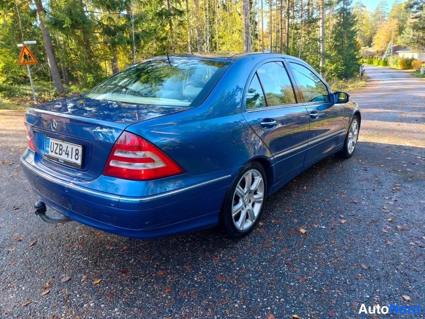 Takaviisto: Sinisen Mercedes-Benzin takaosan kolmeneljänneskuva vasemmalta paikallaan pihatiella, rekisterikilpi, taustalla metsaa.