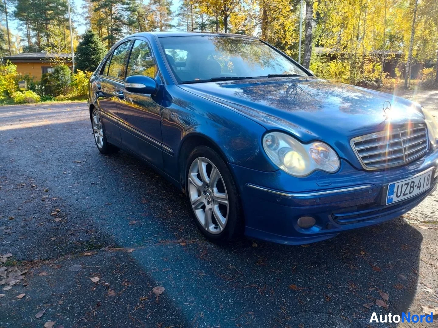 Etuviisto, nokka oikealle: Sinisen Mercedes-Benzin etuosan kolmeneljänneskuva paikallaan maantiella, etulokasuojissa vahinkoja, rekisterikilpi, taustalla metsaa.