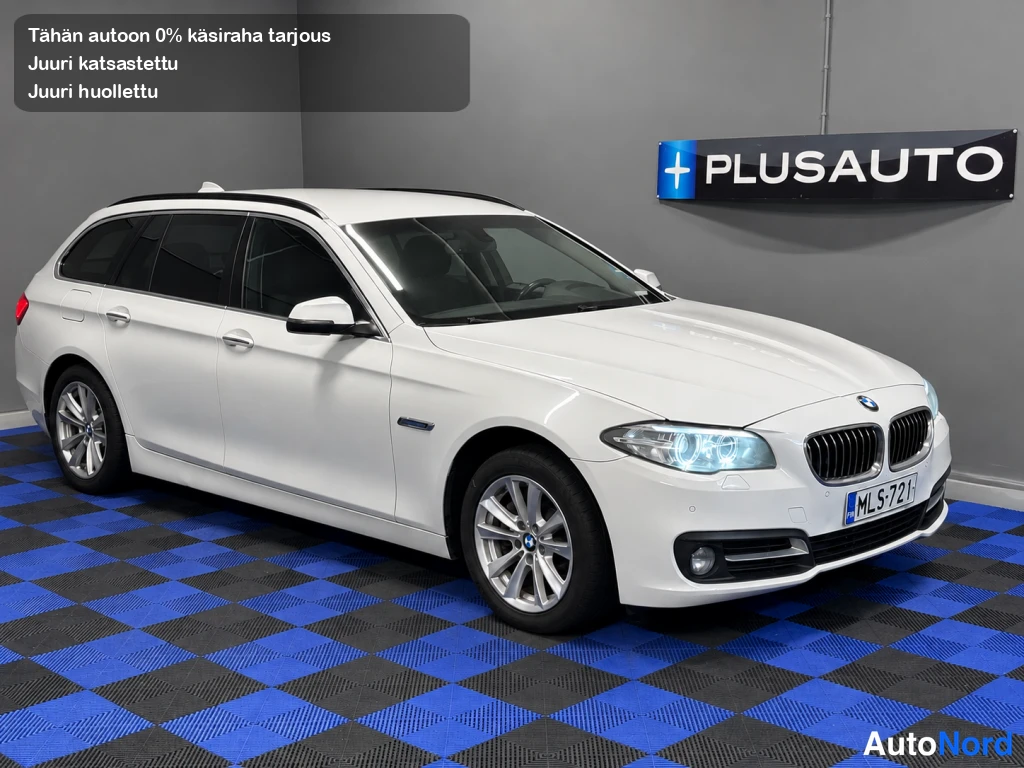 Etuviisto, nokka oikealle: Valkoinen BMW 218d Touring keulakulmasta sisätiloissa, etusäleikkö ja kylki näkyvissä sinimustaruutuisella lattialla.