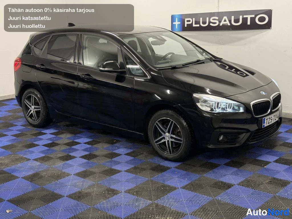 Etuviisto, nokka oikealle: Musta BMW 218d Touring kylki- ja keulakulmasta sisätiloissa sinimustaruutuisella lattialla, etusäleikkö ja etupyörä näkyvät.