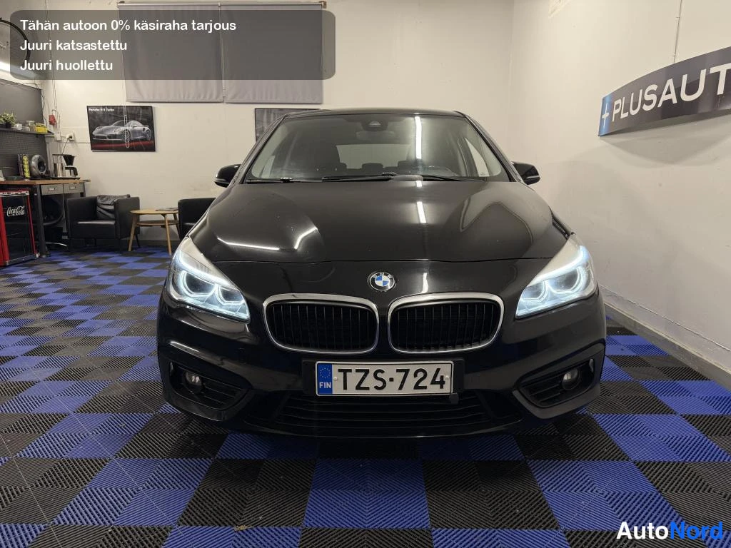 Edestä: Mustan BMW 218d:n keulakuva sisätiloissa, rekisterikilpi ja PLUSAUTO-kyltti taustalla.