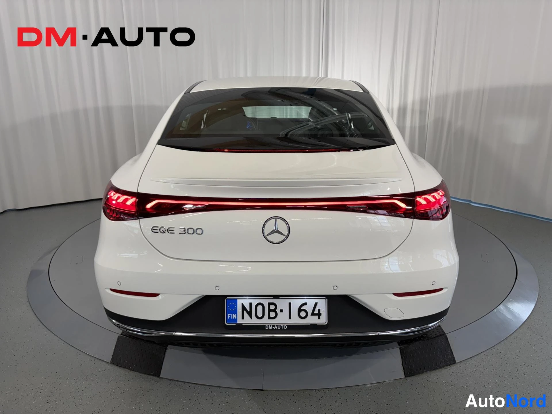 Takaa: Valkoisen Mercedes-Benz EQE 300:n takaosa suoraan takaa: punainen kevytvalo juova konepellin reunassa, EQE 300 -merkintä, Mercedes-tähti ja rekisterikilpi.