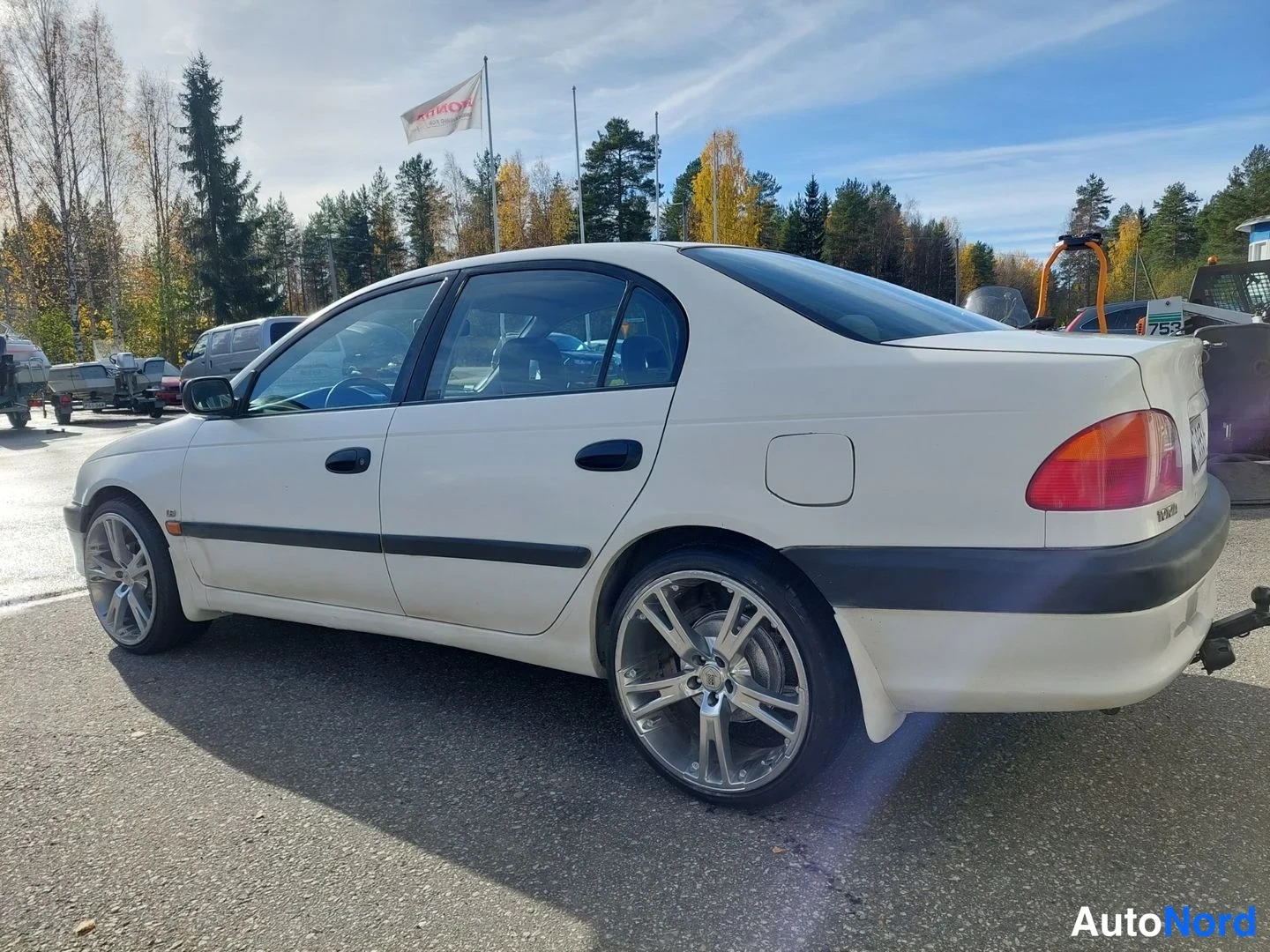 Takaviisto: Valkoinen Toyota Avensis -sedan kolmeneljäsosakulmasta takaa vasemmalle, neliovinen korimalli ja hopeavanteet näkyvissä.