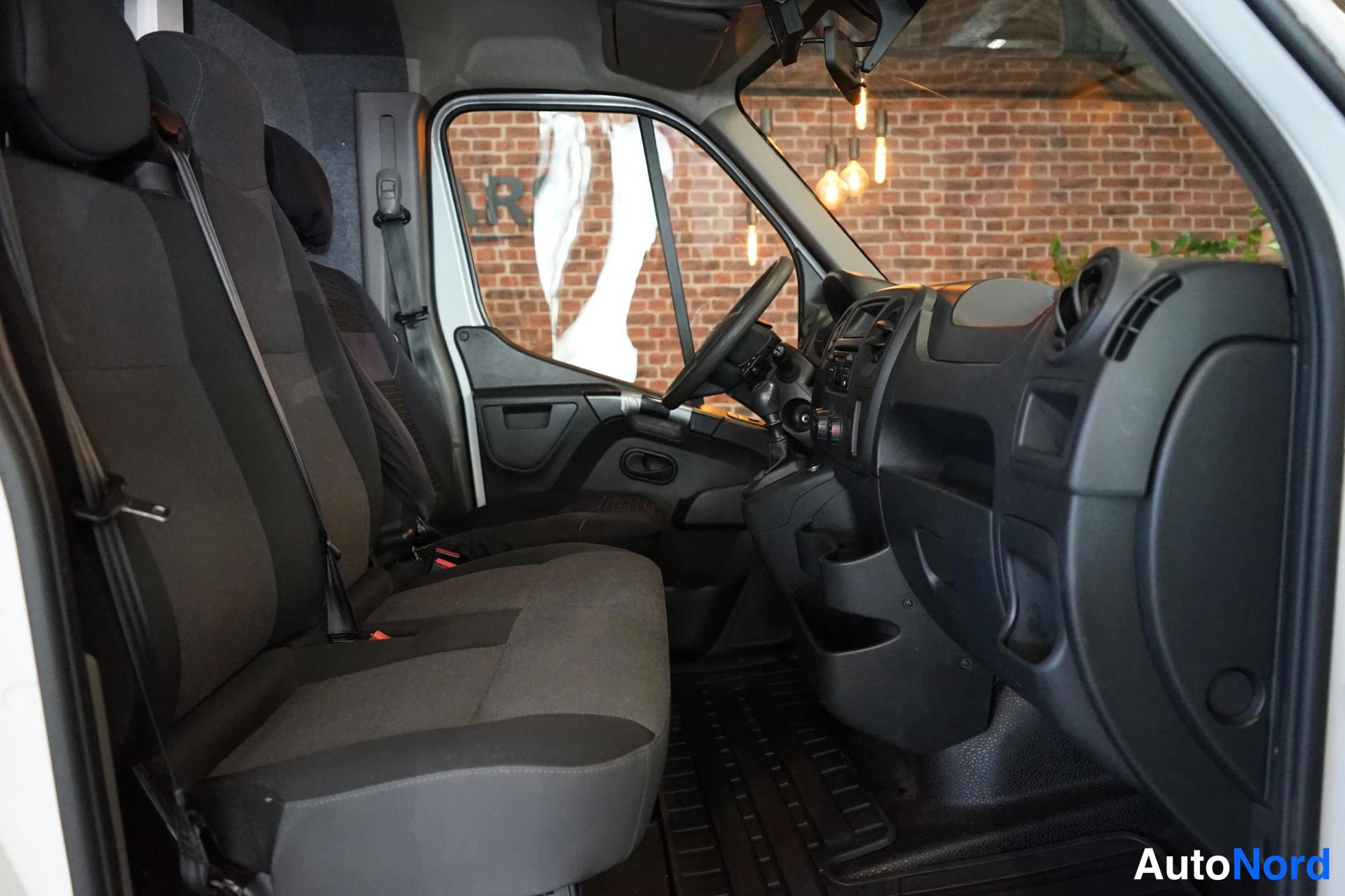Muu: Nissan NV400:n sisätilat takaovelta nähtynä: kaksi istuinrivejä tummin kankaisin, turvavyöt ja tummat muovipinnat kojelaudassa; taustalla punatiilinen seinä.