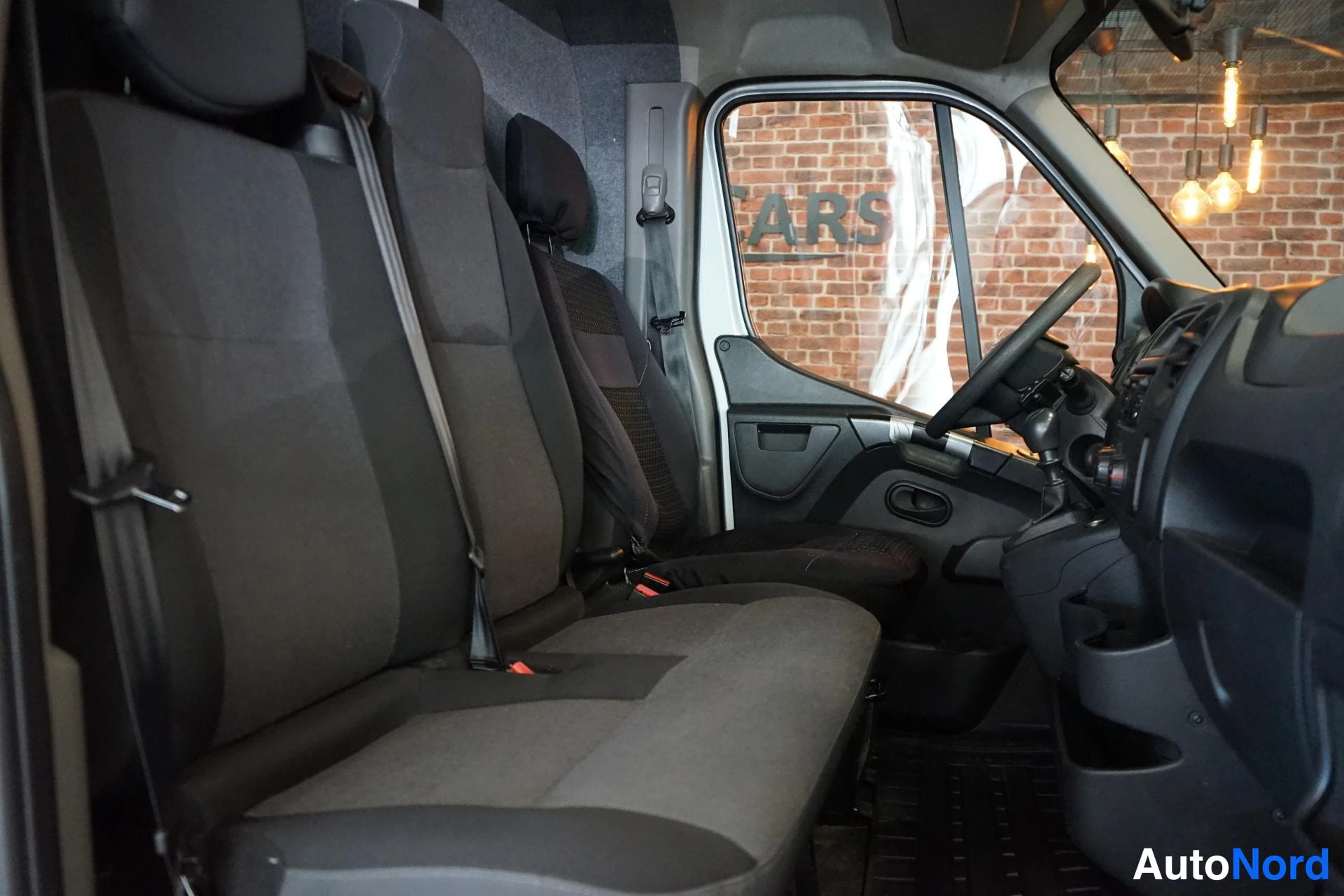 Muu: Nissan NV400:n matkustamotilat takaovelta nähtynä: tummat kankaiset istuimet, turvavyöt, kojelauta ja ohjauspyörä oikealla; taustalla tiiliseinä.