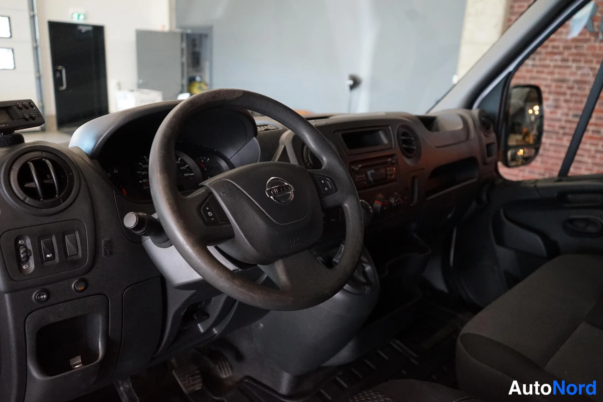 Muu: Ohjaamon sisäkuva: harmaa Nissan-ohjauspyörä, mittaristo ja tumma kojelauta.