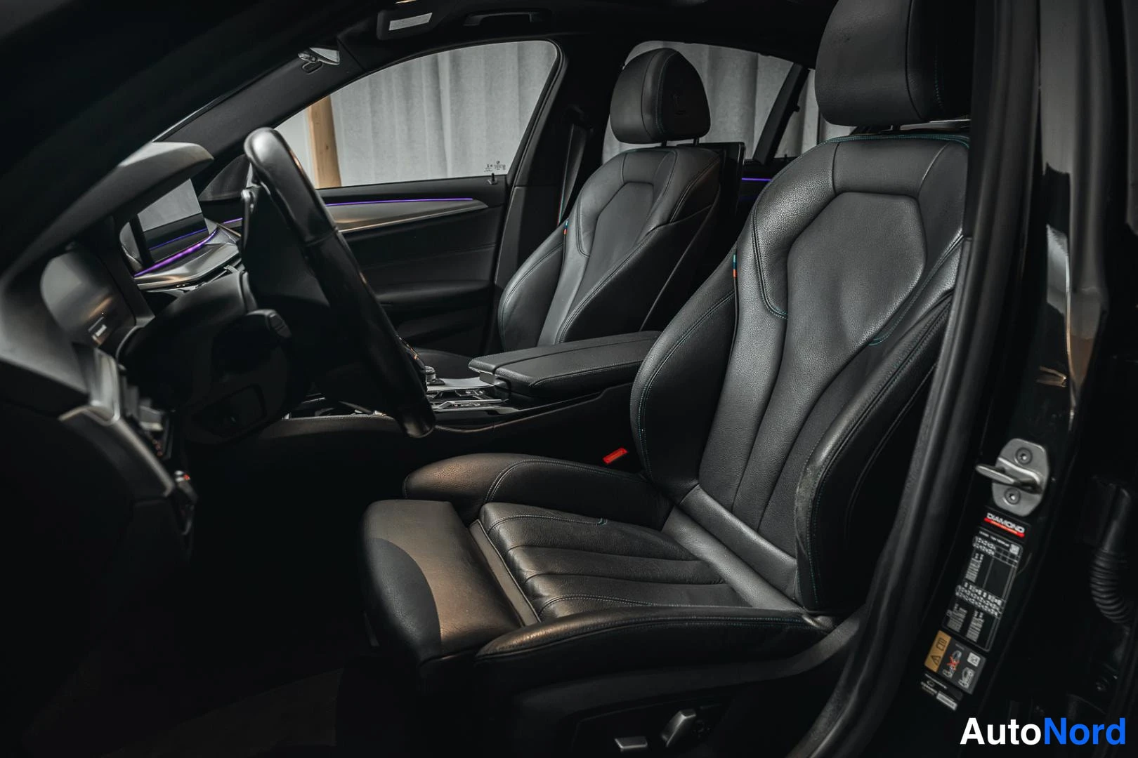 Yksityiskohta: Interno di un'auto con sedili in pelle sportivi neri, volante e cruscotto illuminato da luce d'ambiente; porta aperta con etichetta informativa sul montante.