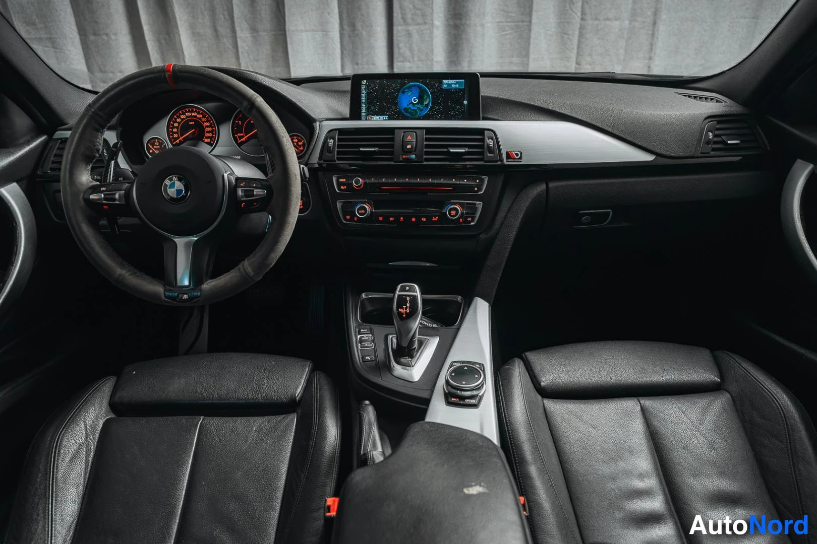 Muu: Auton etuistuimilta kuvattu näkymä ohjaamoon: BMW-ohjauspyörä, mittaristo, keskikonsolilla infotainment-näyttö ja automaattivaihteen valitsin, mustat nahkaistui