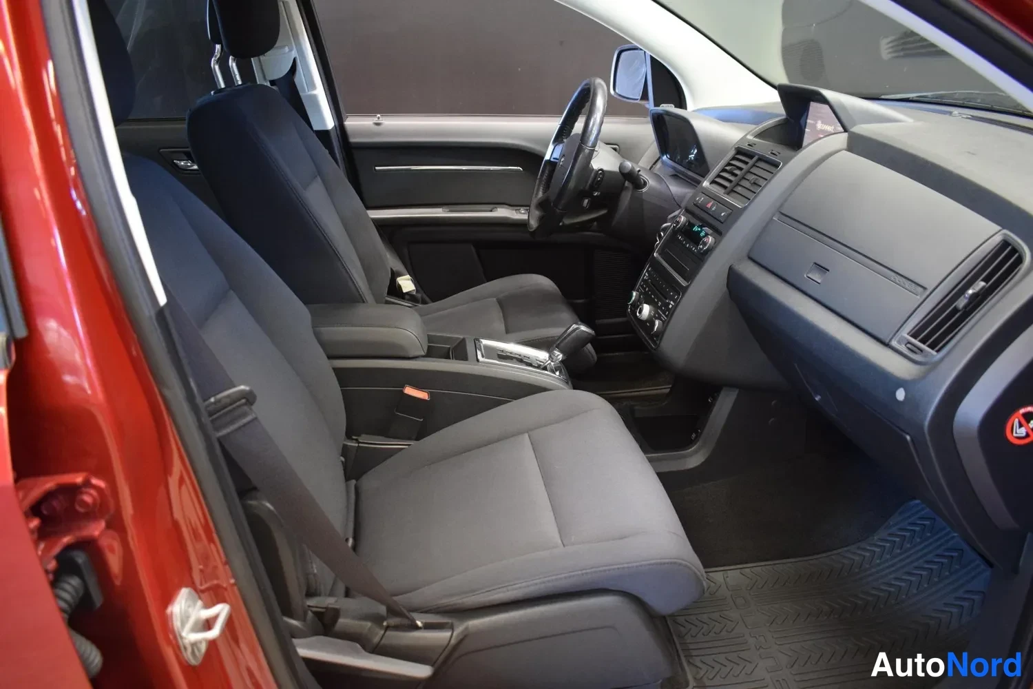 Sivulta: Dodge Journey -henkilöautin sisätila: kaksi tummaa etuistuinta, ohjauspyörä, automaattivaihteen valitsin, kojelauta ja kumimatot näkyvät avatulta kuljettajan pu